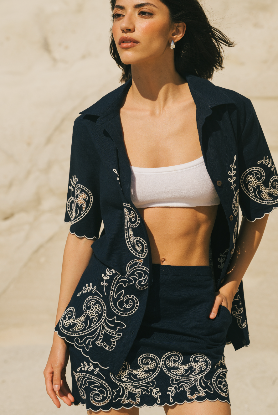 Woman wearing a dark blue embroidered jacket over a white top and black shorts on a beige background Calling June