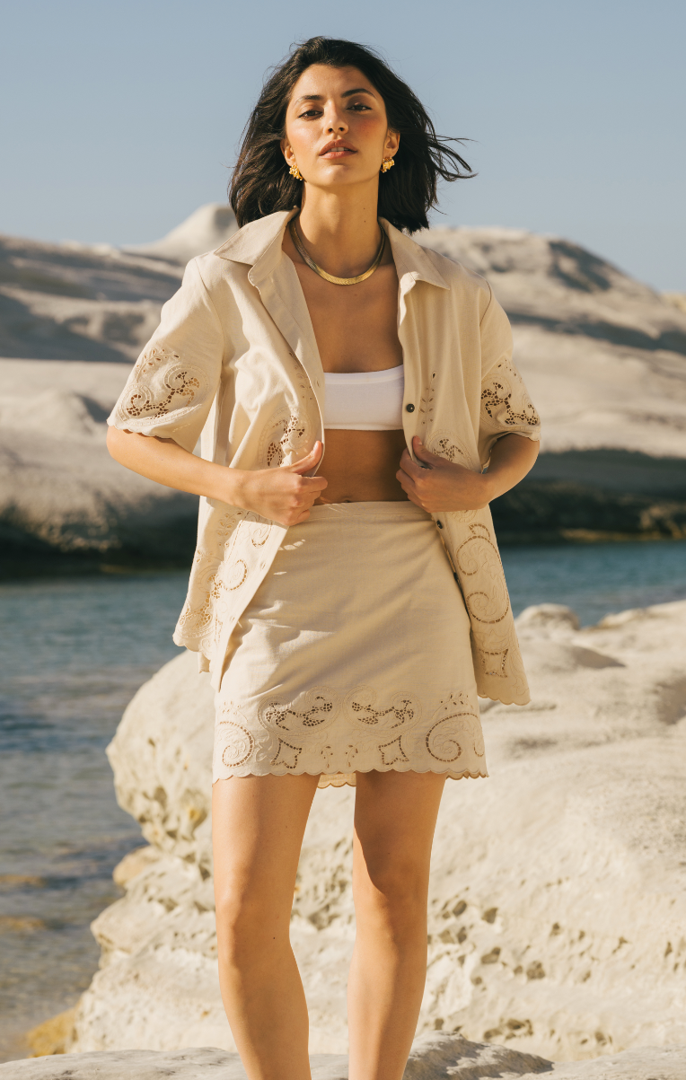 Woman in a beige outfit standing on a rocky beach with mountains in the background Calling June