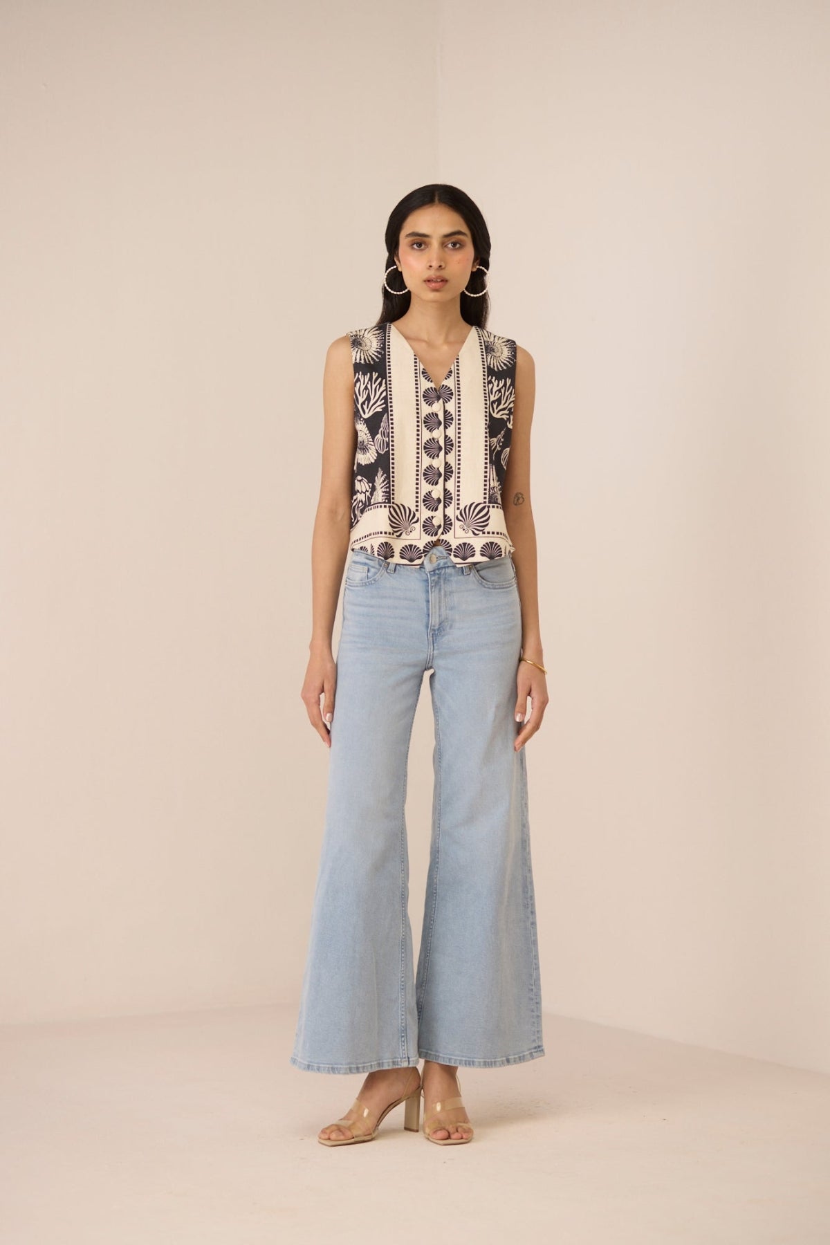Woman wearing a sleeveless top with floral embroidery and light blue jeans on a beige background Calling June