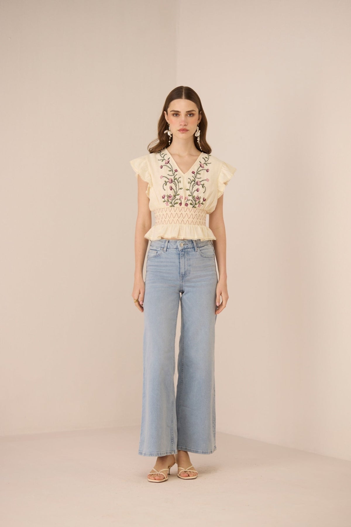 Woman wearing a beige embroidered top and light blue jeans on a beige background Calling June