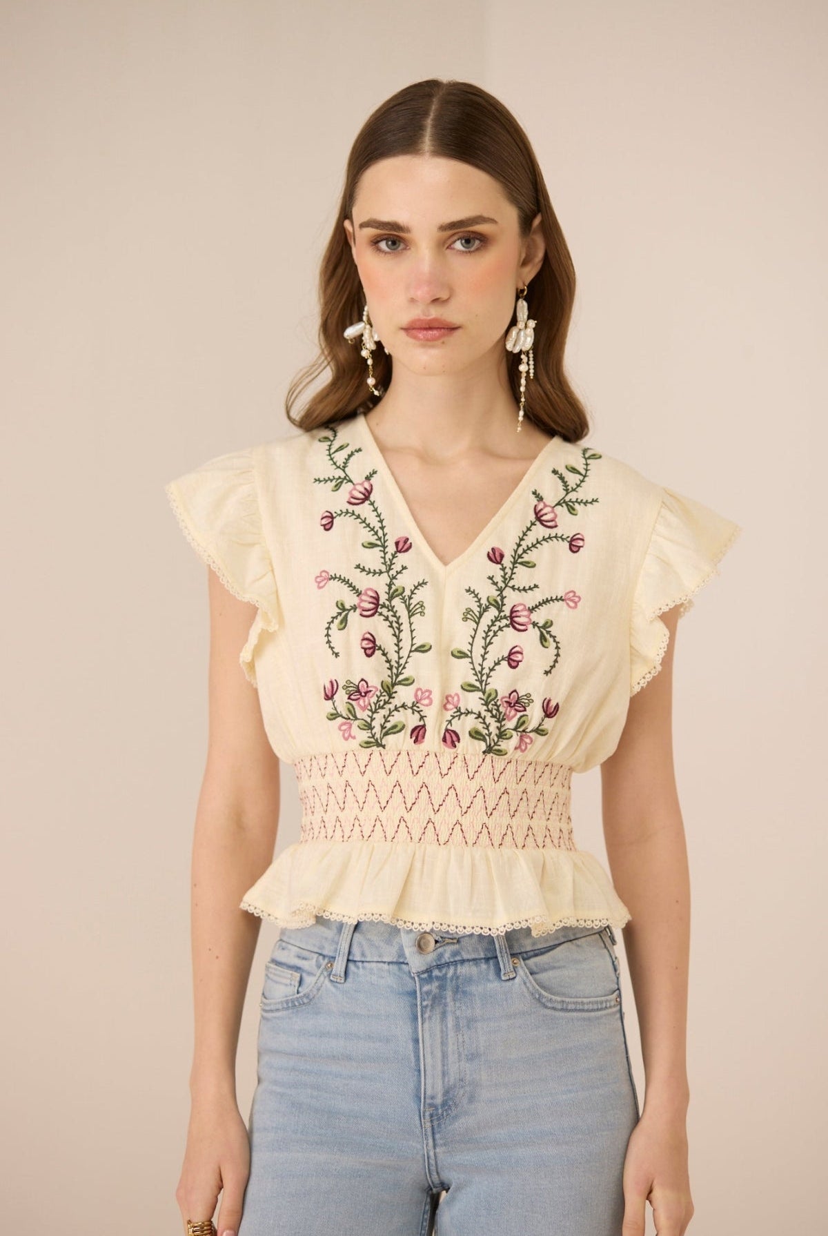 Woman wearing a beige top with floral embroidery and light blue jeans against a beige background Calling June