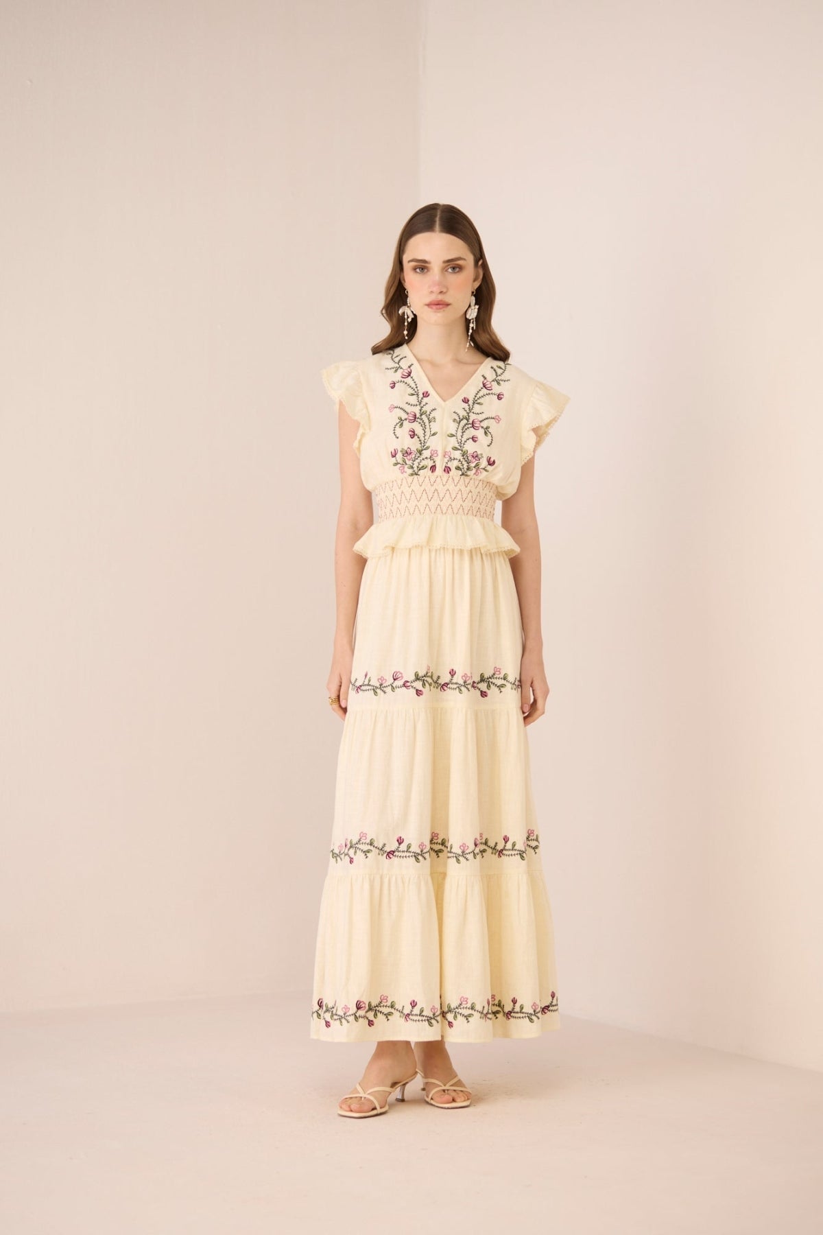 Woman wearing a long, light-colored dress with floral embroidery on a plain background Calling June