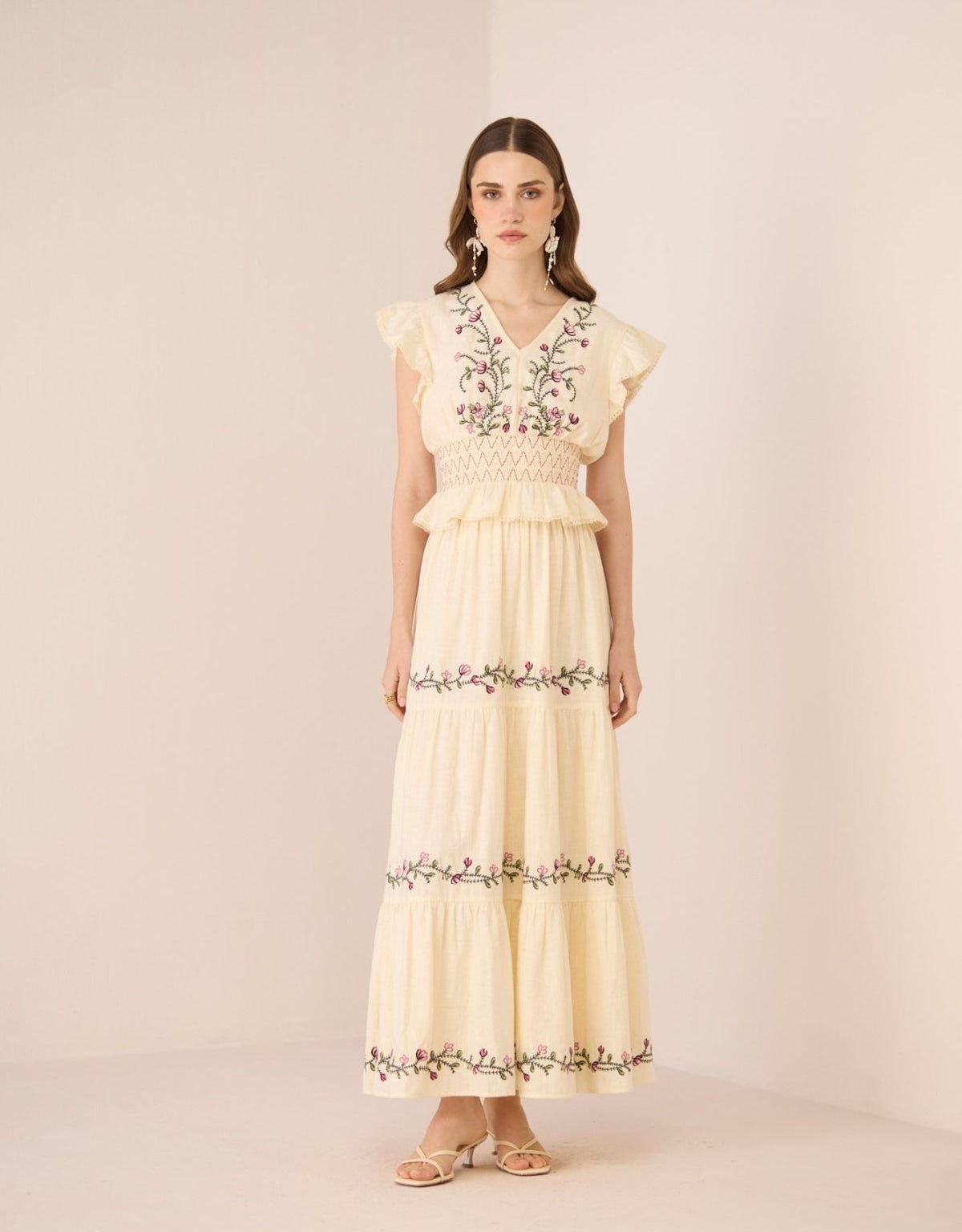 Woman wearing a long, light-colored dress with floral embroidery on a plain background Calling June