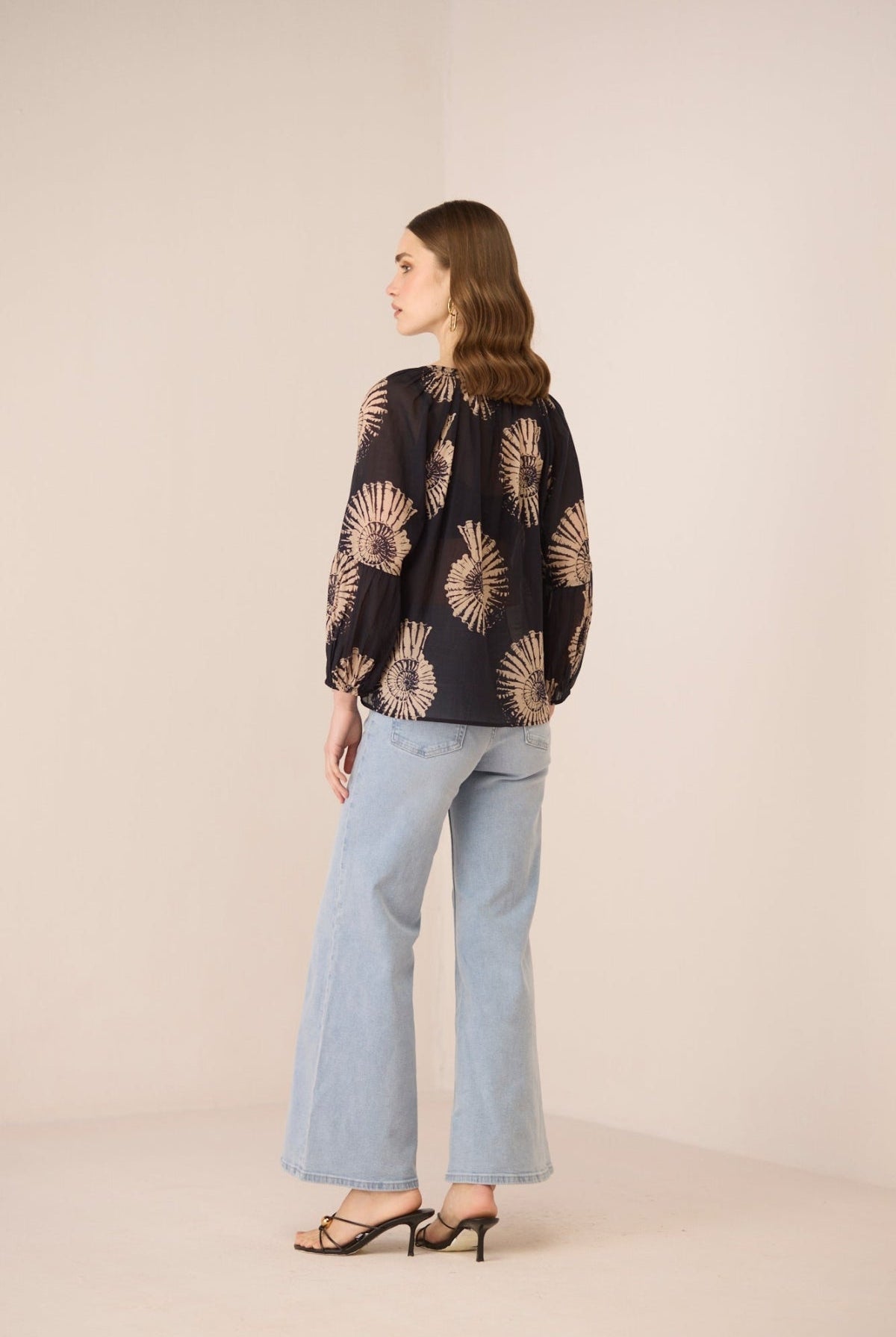 Woman wearing a black blouse with gold patterns and light blue jeans on a beige background Calling June