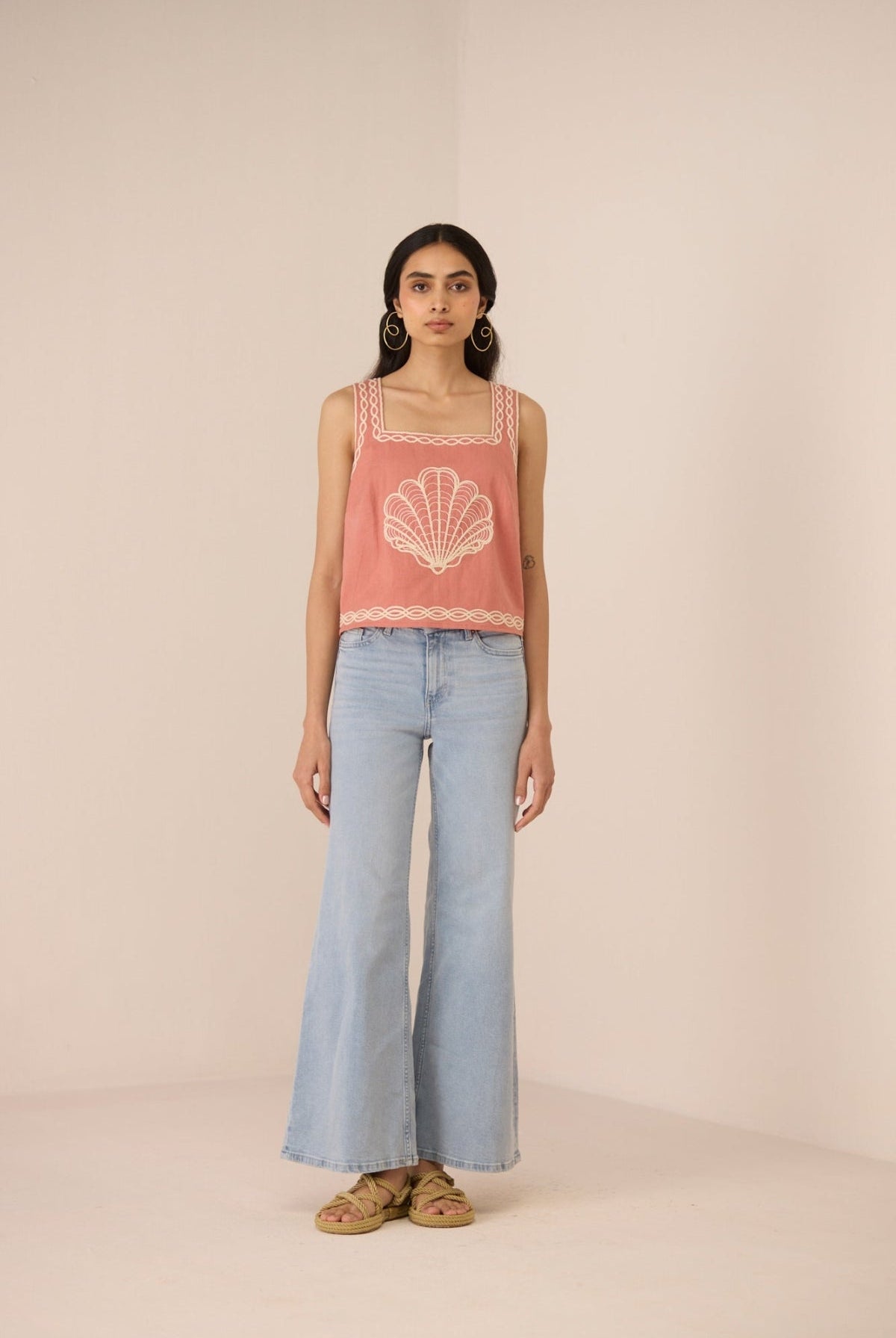 Woman wearing a pink sleeveless top with a shell design and light blue jeans on a beige background Calling June