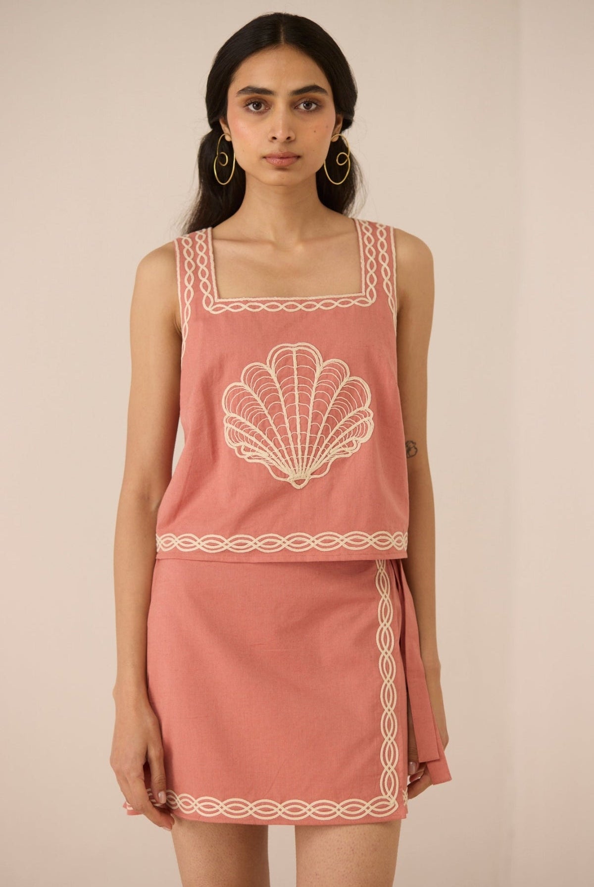 Woman wearing a pink sleeveless top and skirt with white embroidery on a beige background Calling June