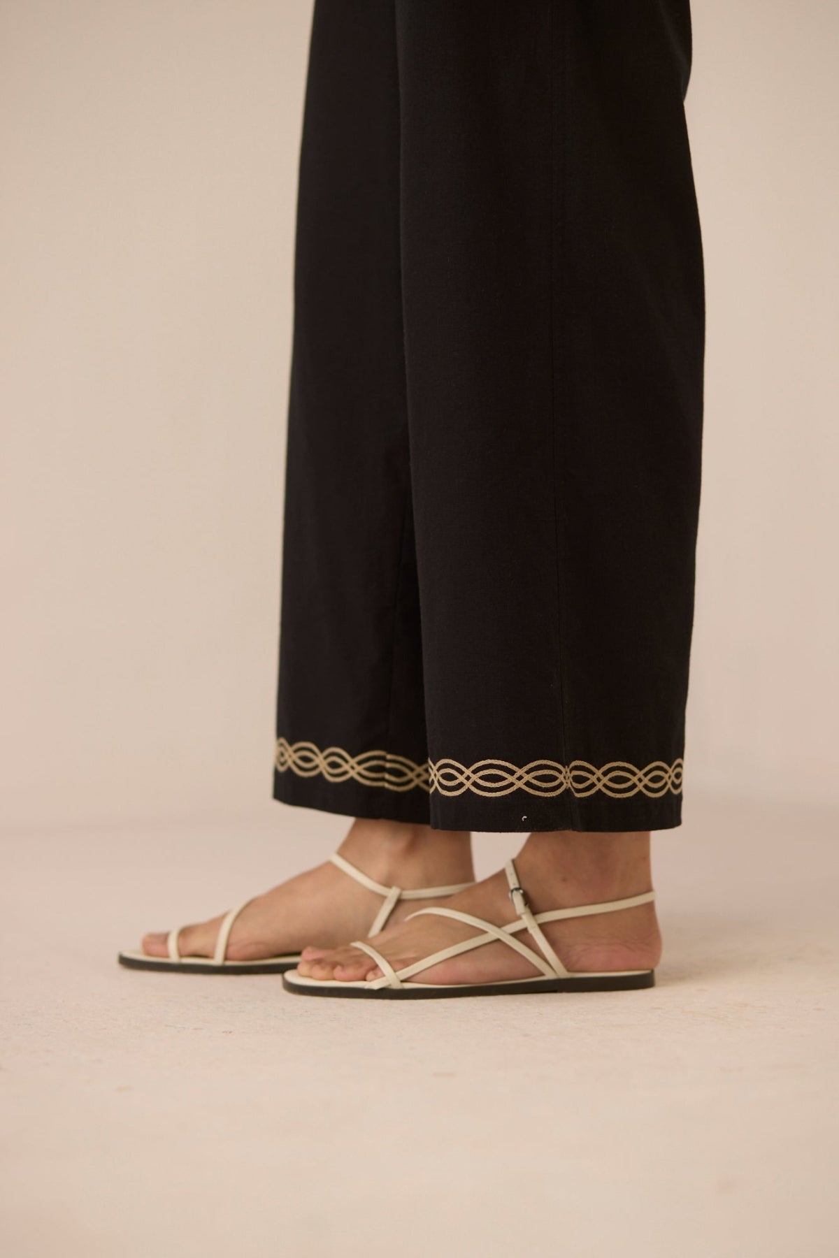 Person wearing black pants with gold embroidery and beige sandals on a beige background Calling June