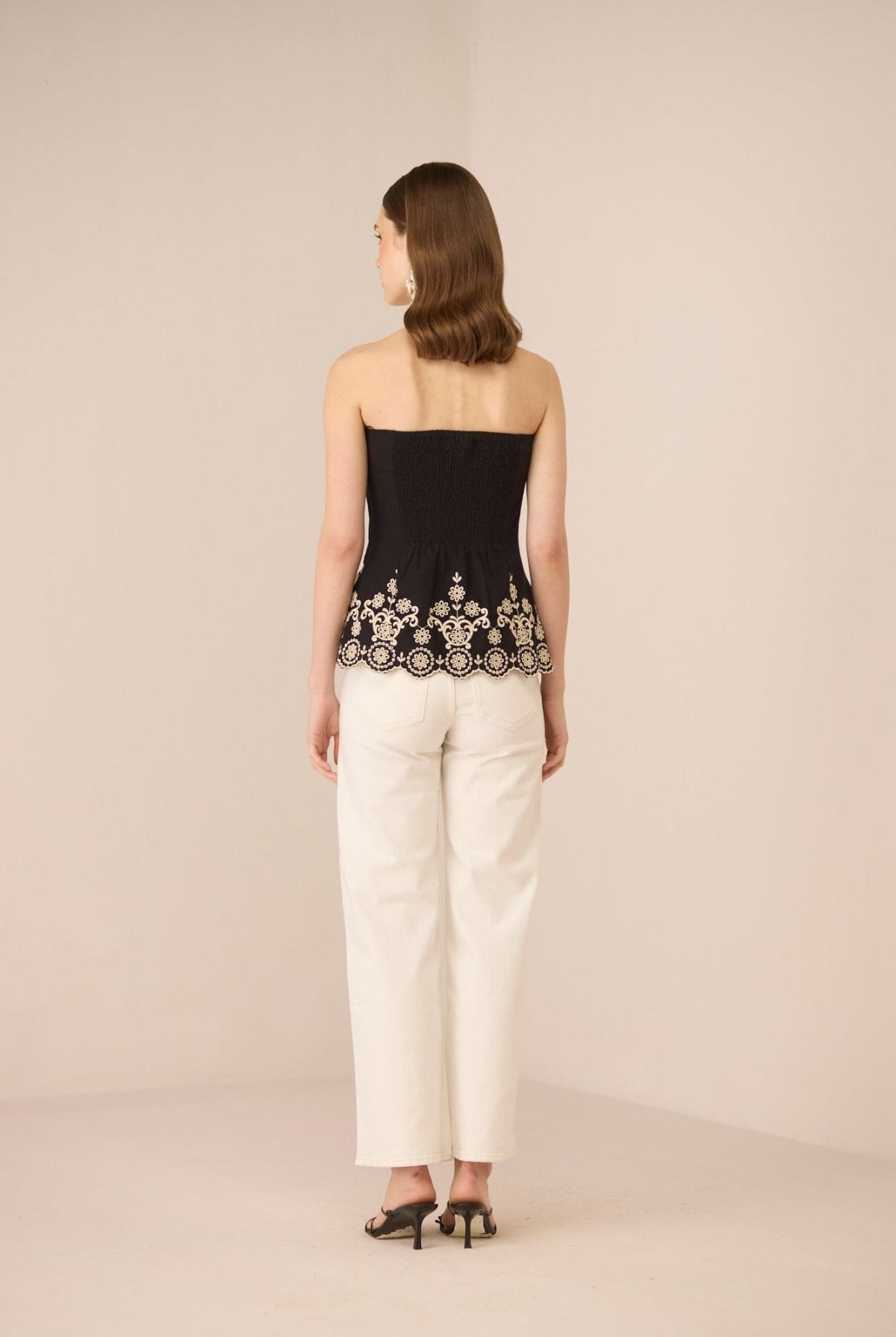 Woman wearing a black strapless top with lace detailing and white pants on a beige background Calling June