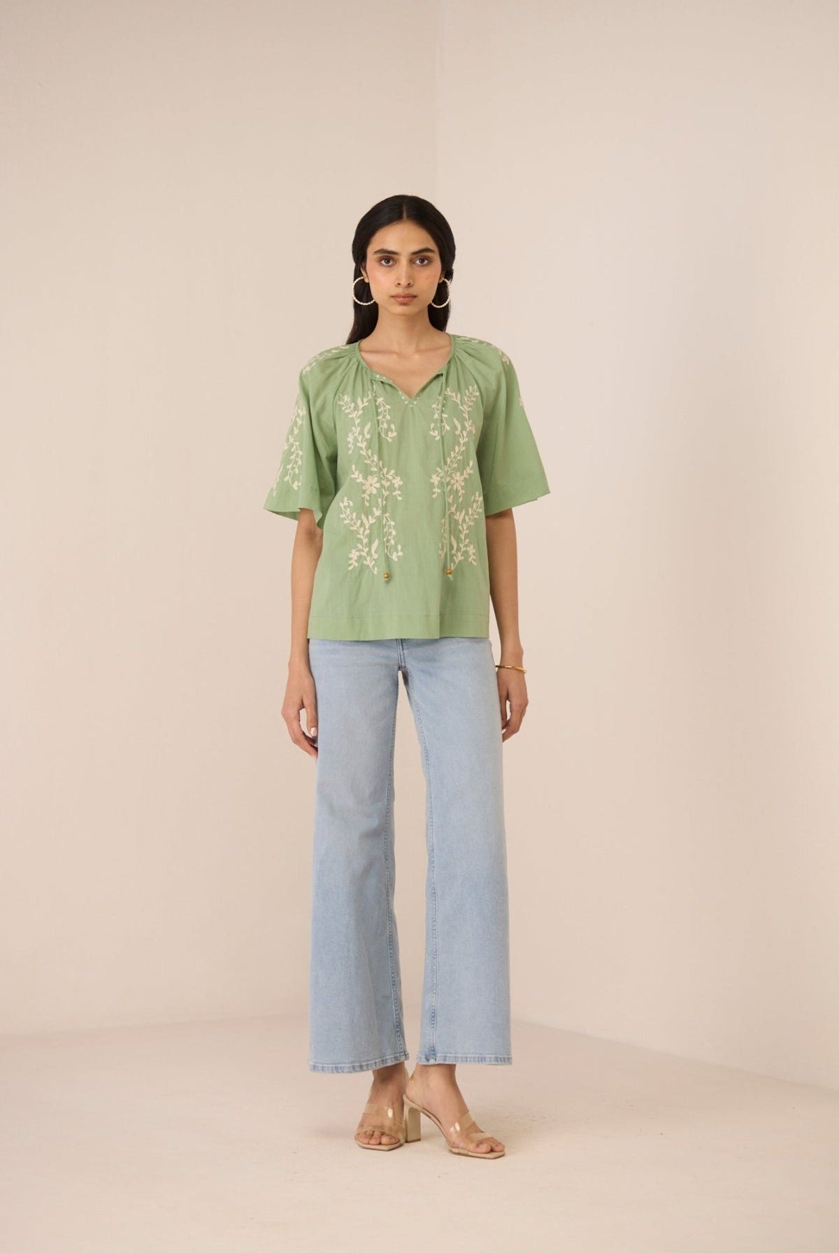 Woman wearing a green embroidered top and light blue jeans on a beige background Calling June