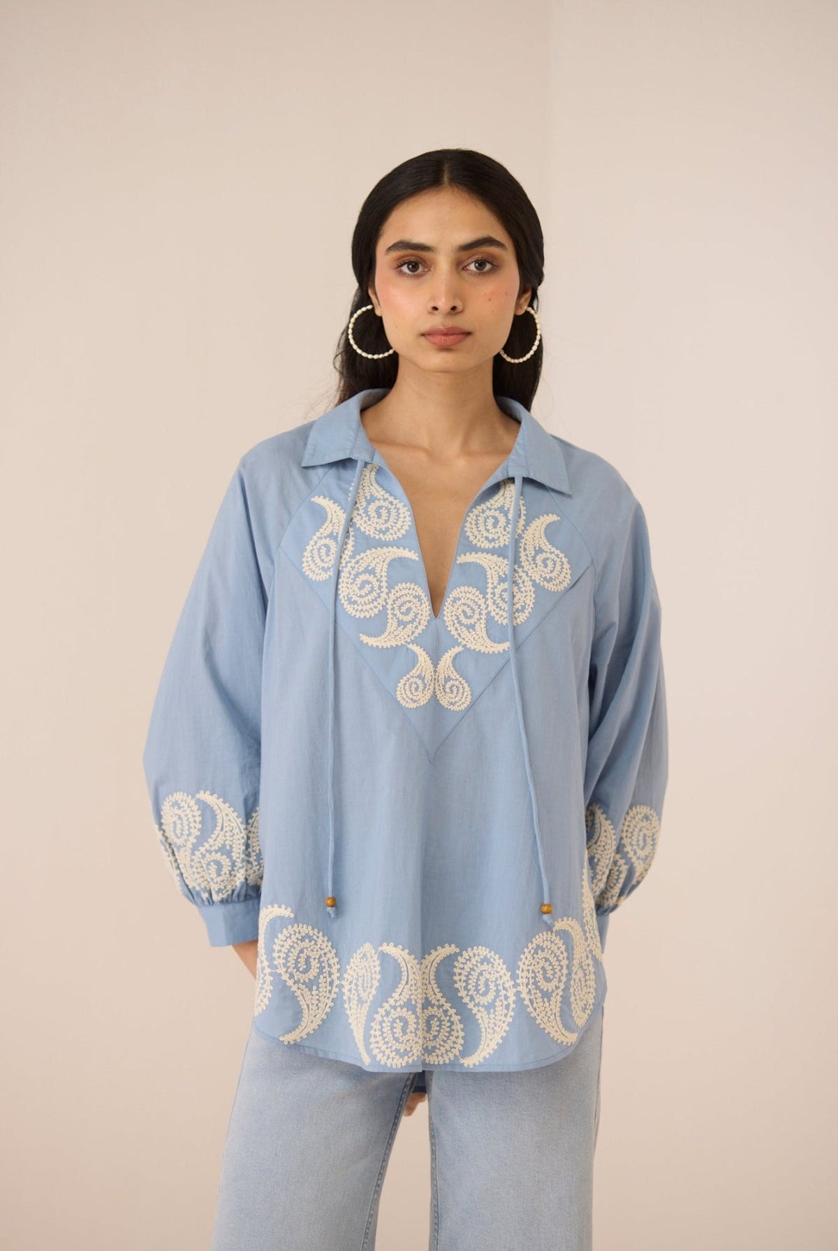 Woman wearing a light blue blouse with intricate white embroidery on a beige background Calling June