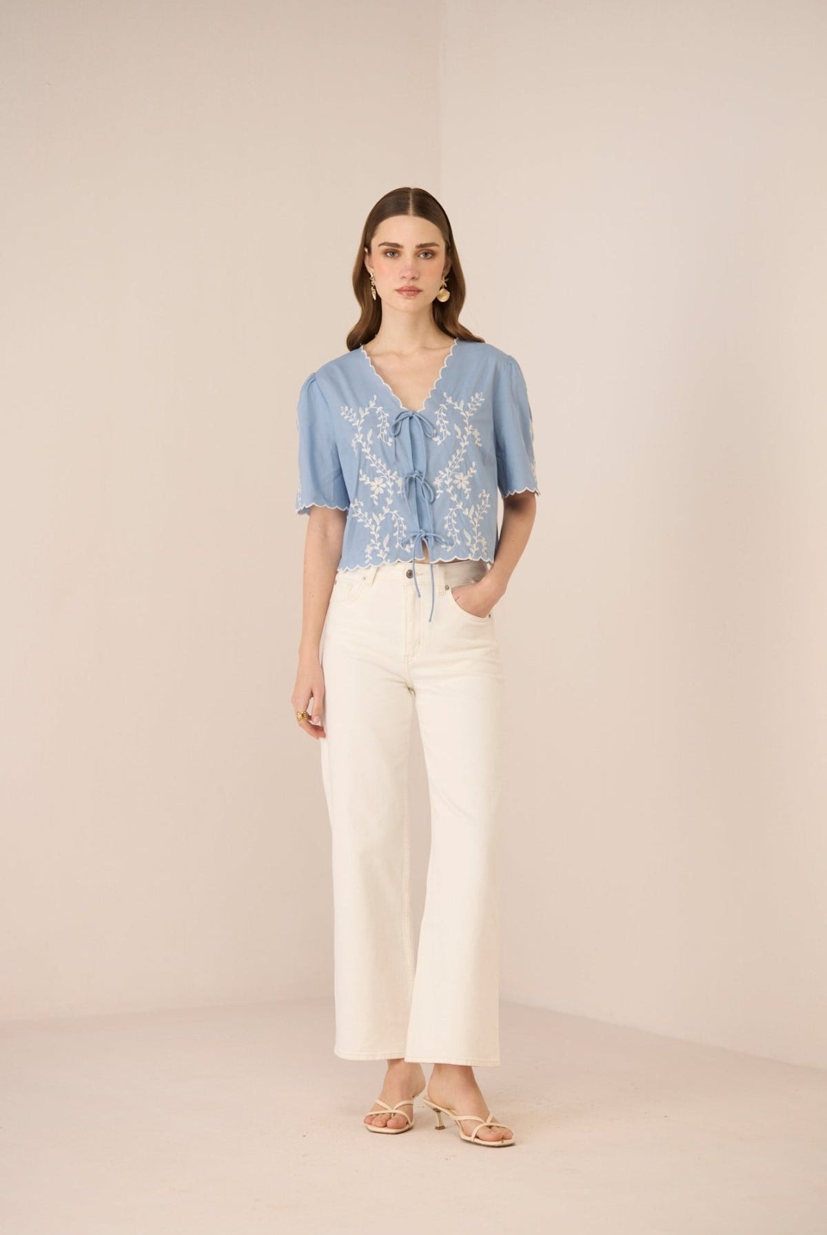 Woman wearing a blue embroidered blouse and white pants on a beige background Calling June