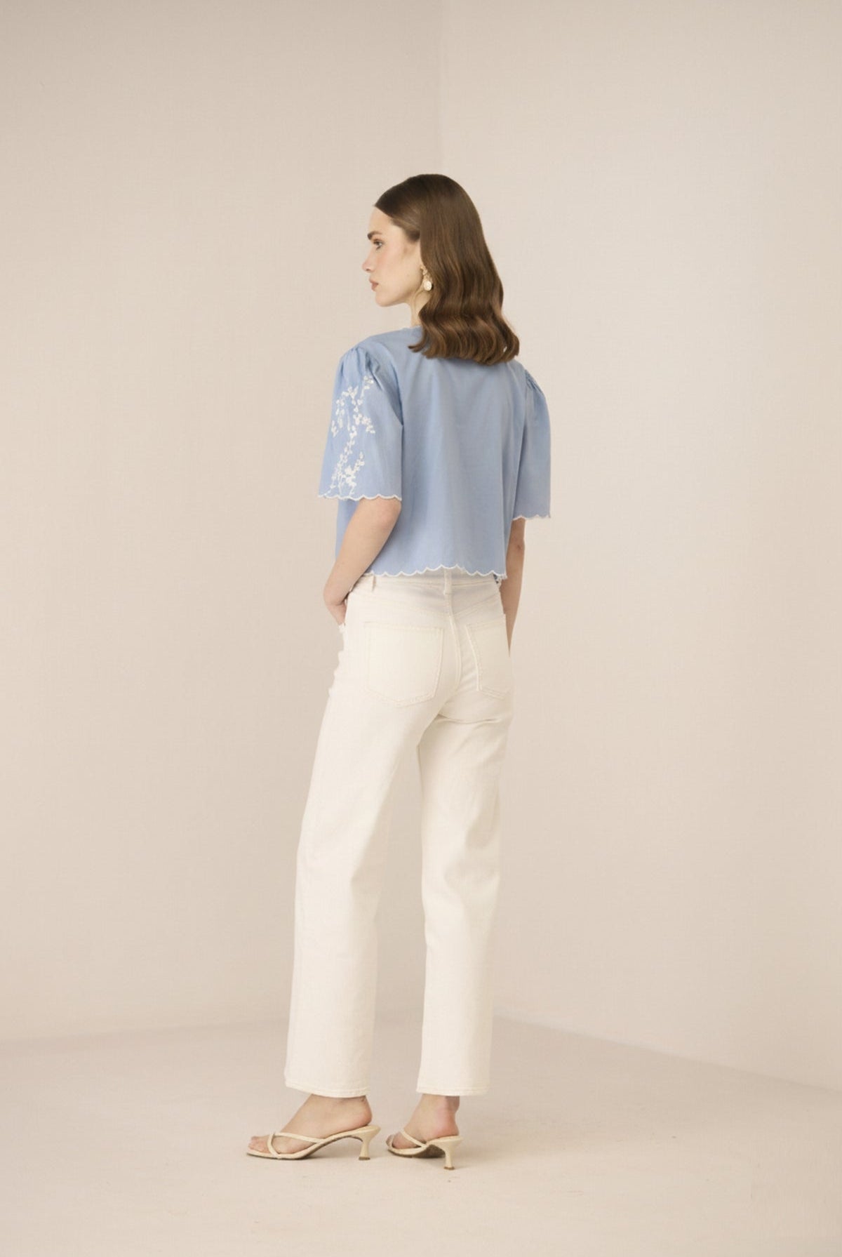 Woman wearing a light blue top with lace sleeves and white pants on a beige background Calling June