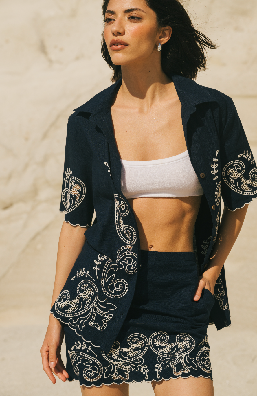 Woman wearing a dark blue embroidered jacket over a white top and black shorts on a beige background Calling June
