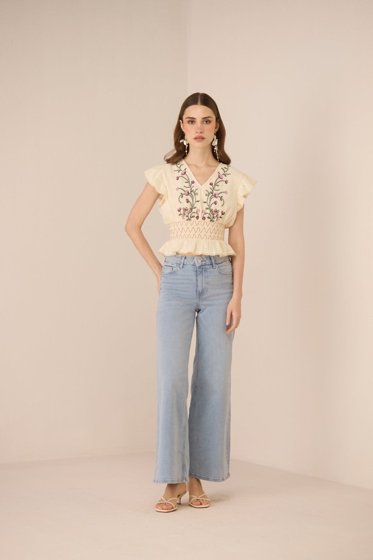 Woman wearing a beige embroidered top and light blue jeans on a beige background Calling June