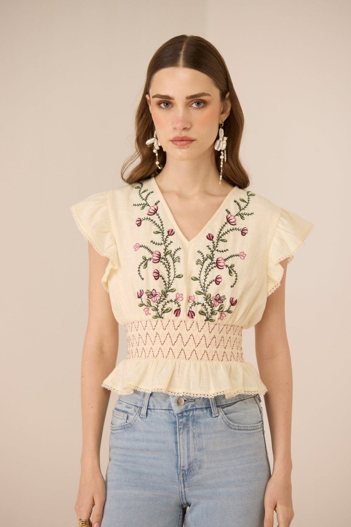 Woman wearing a beige top with floral embroidery and light blue jeans against a beige background Calling June