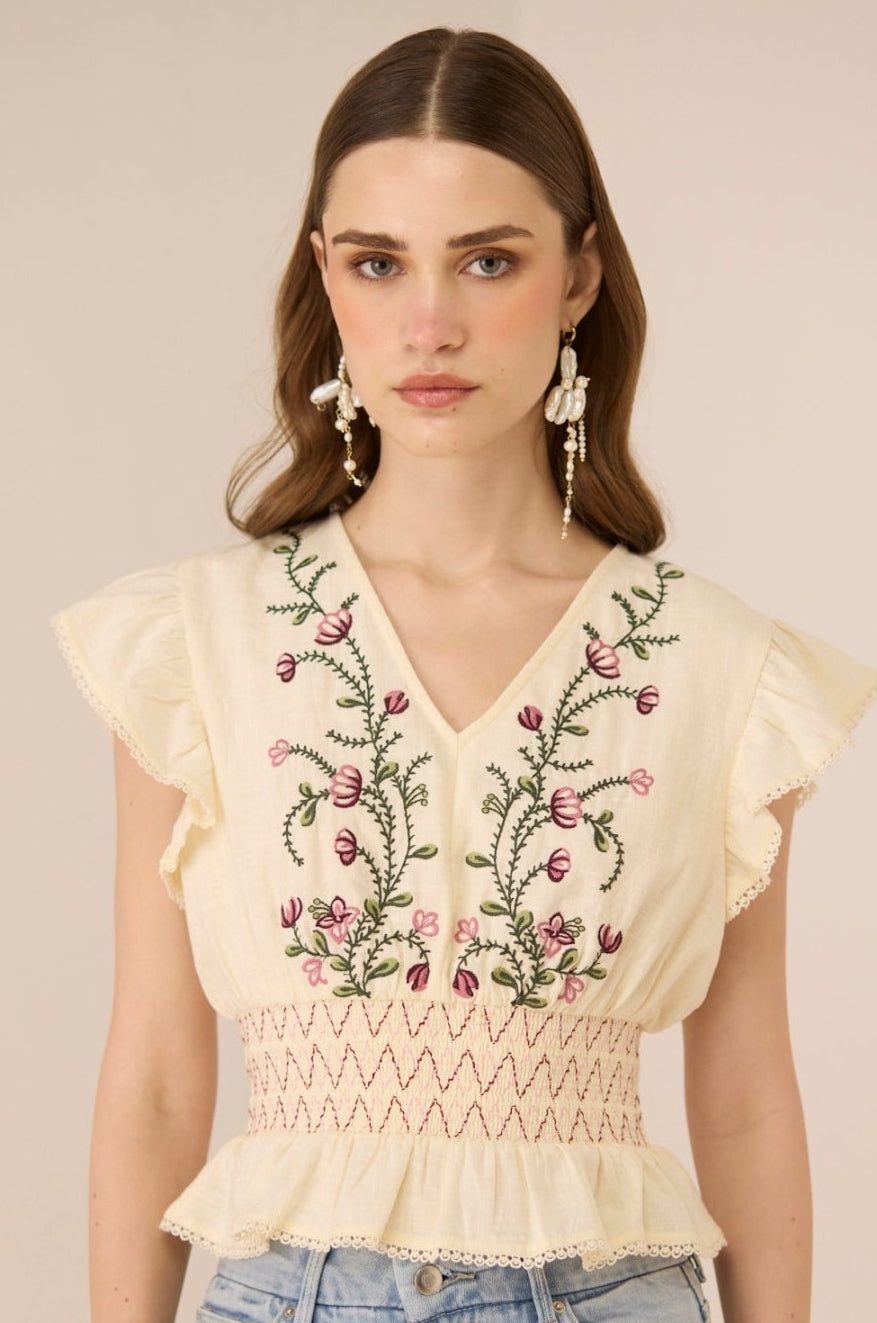 Woman wearing a beige top with floral embroidery and light blue jeans against a beige background Calling June