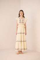 Woman wearing a long, light-colored dress with floral embroidery on a plain background Calling June