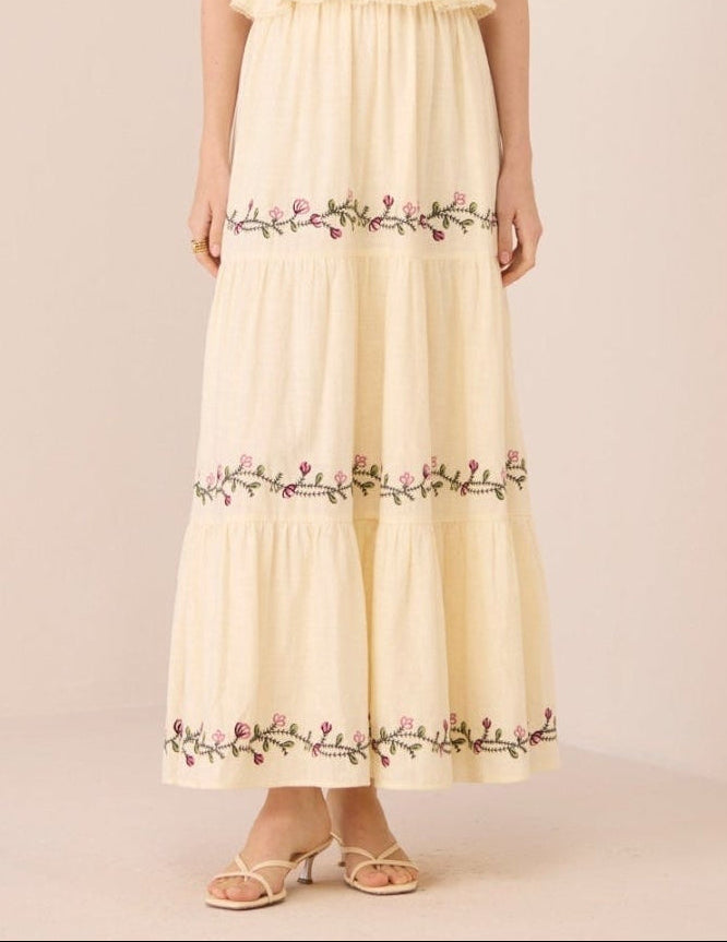 Woman wearing a long, light-colored dress with floral embroidery on a plain background Calling June
