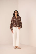 Woman wearing a patterned blouse and white pants against a plain background Calling June