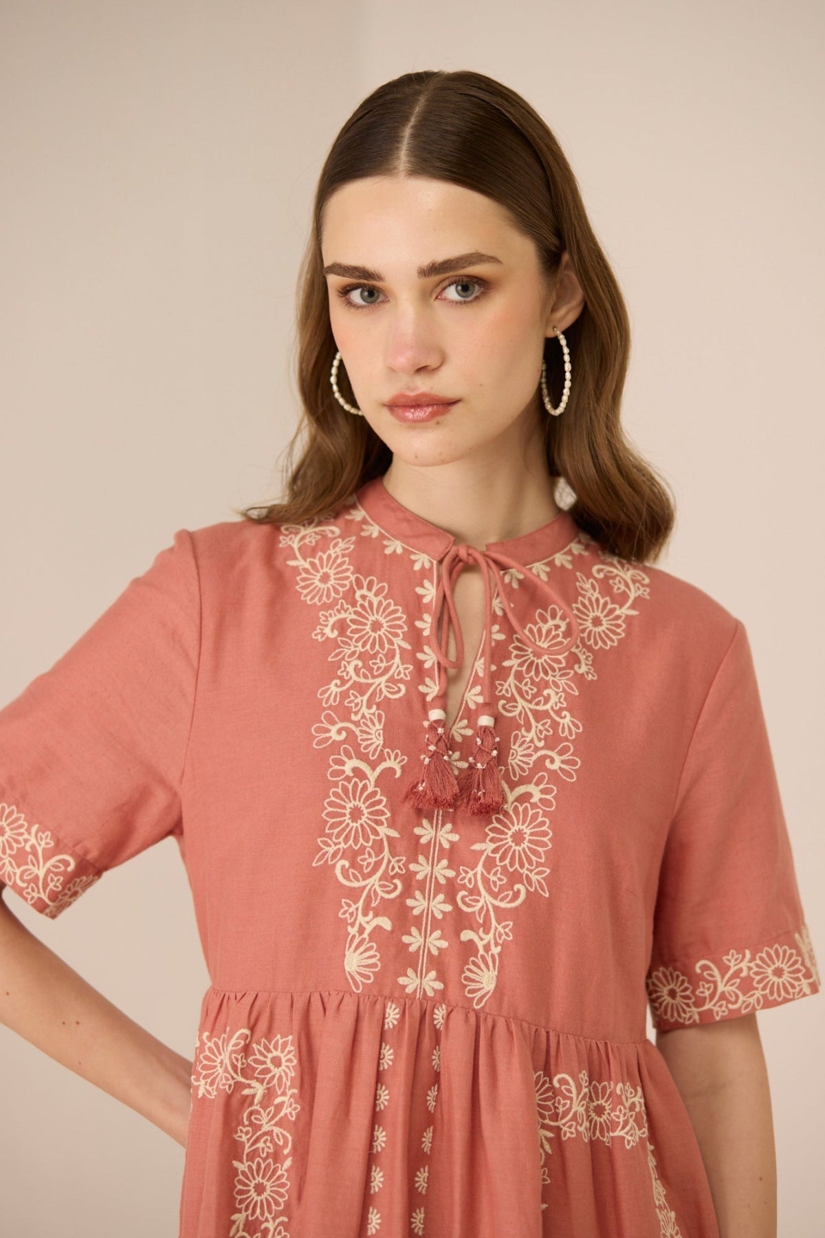 Woman wearing a pink dress with white floral embroidery against a beige background Calling June