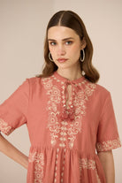 Woman wearing a pink dress with white floral embroidery against a beige background Calling June