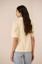 Woman wearing a light yellow polo shirt with floral embroidery on the sleeves against a plain background Calling June