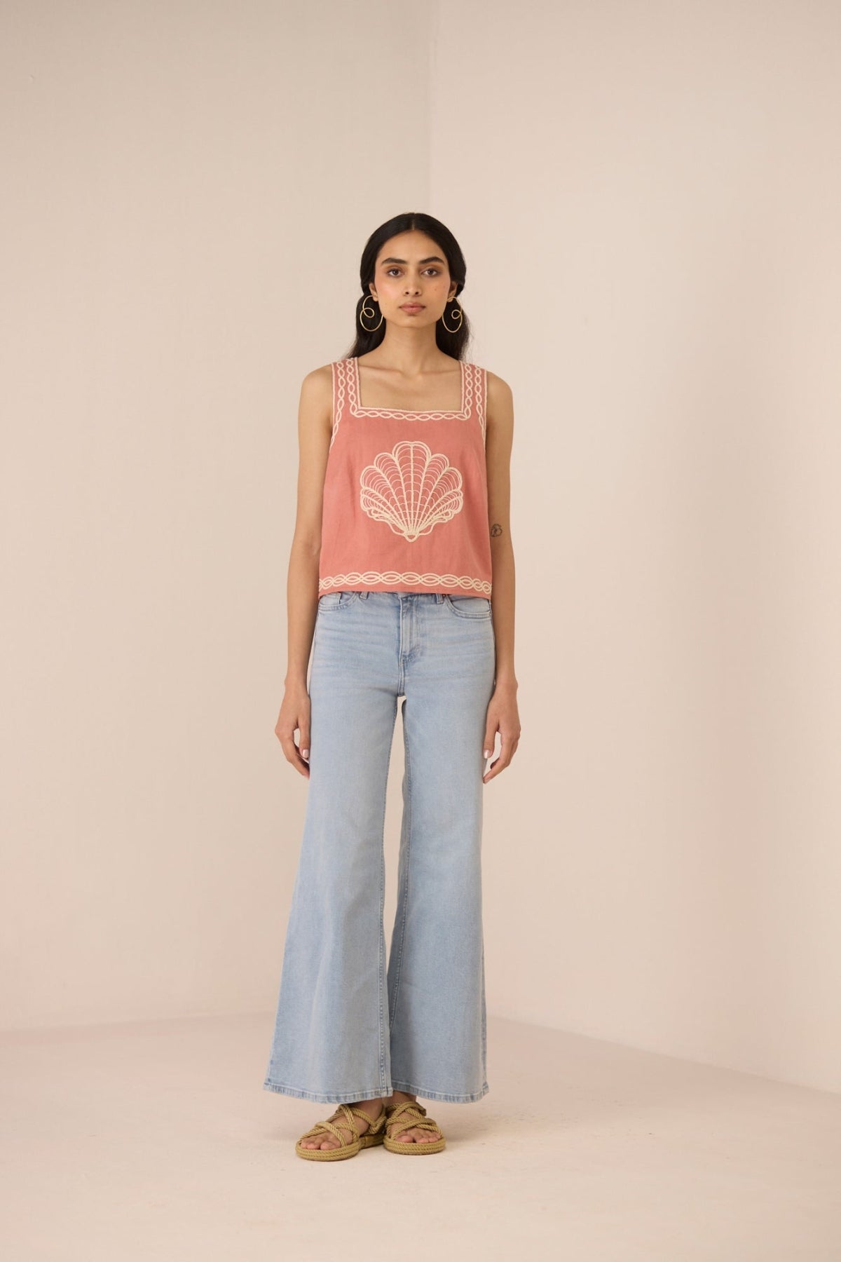 Woman wearing a pink sleeveless top with a shell design and light blue jeans on a beige background Calling June