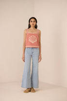 Woman wearing a pink sleeveless top with a shell design and light blue jeans on a beige background Calling June