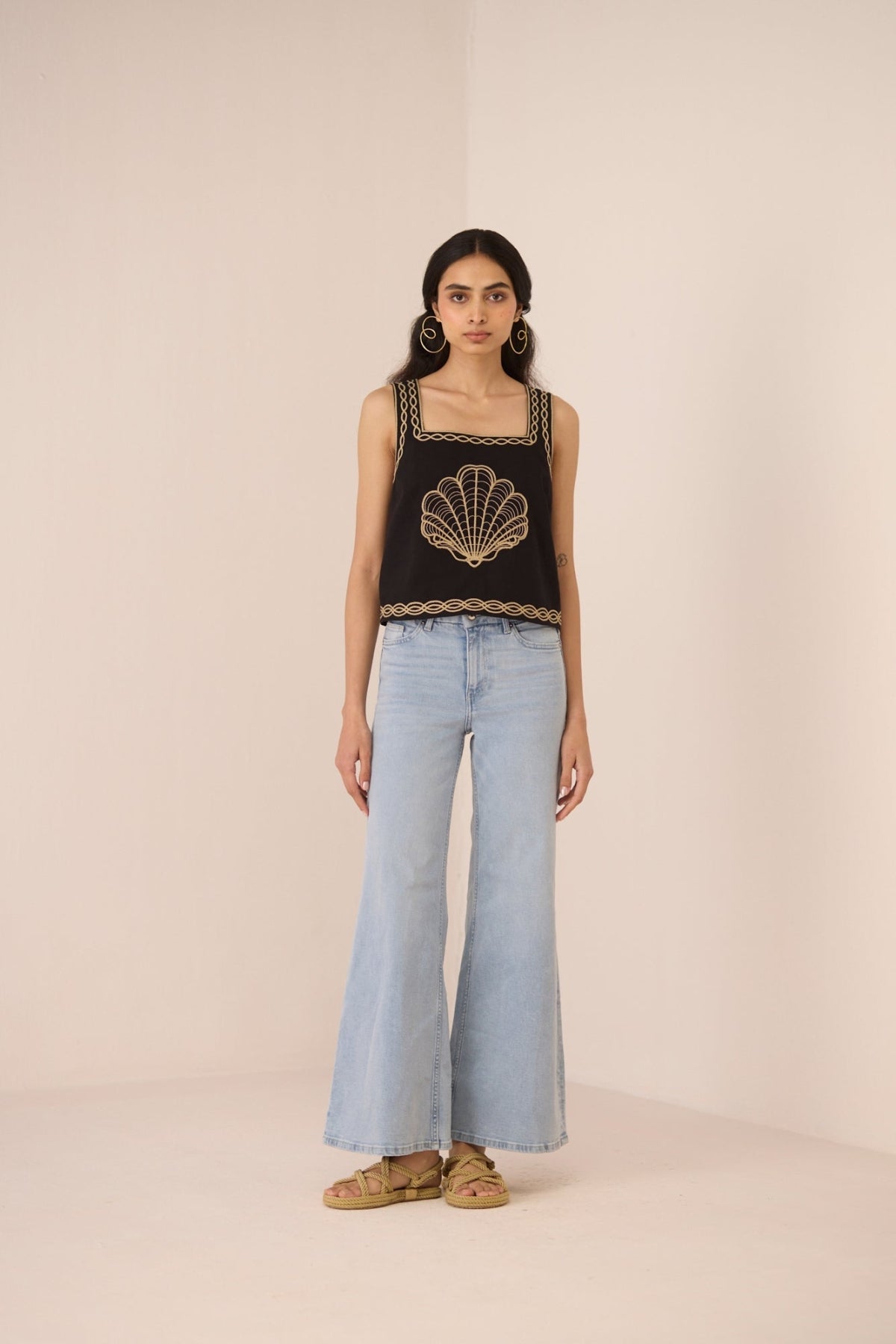 Woman wearing a black sleeveless top with shell design and light blue jeans on a beige background Calling June