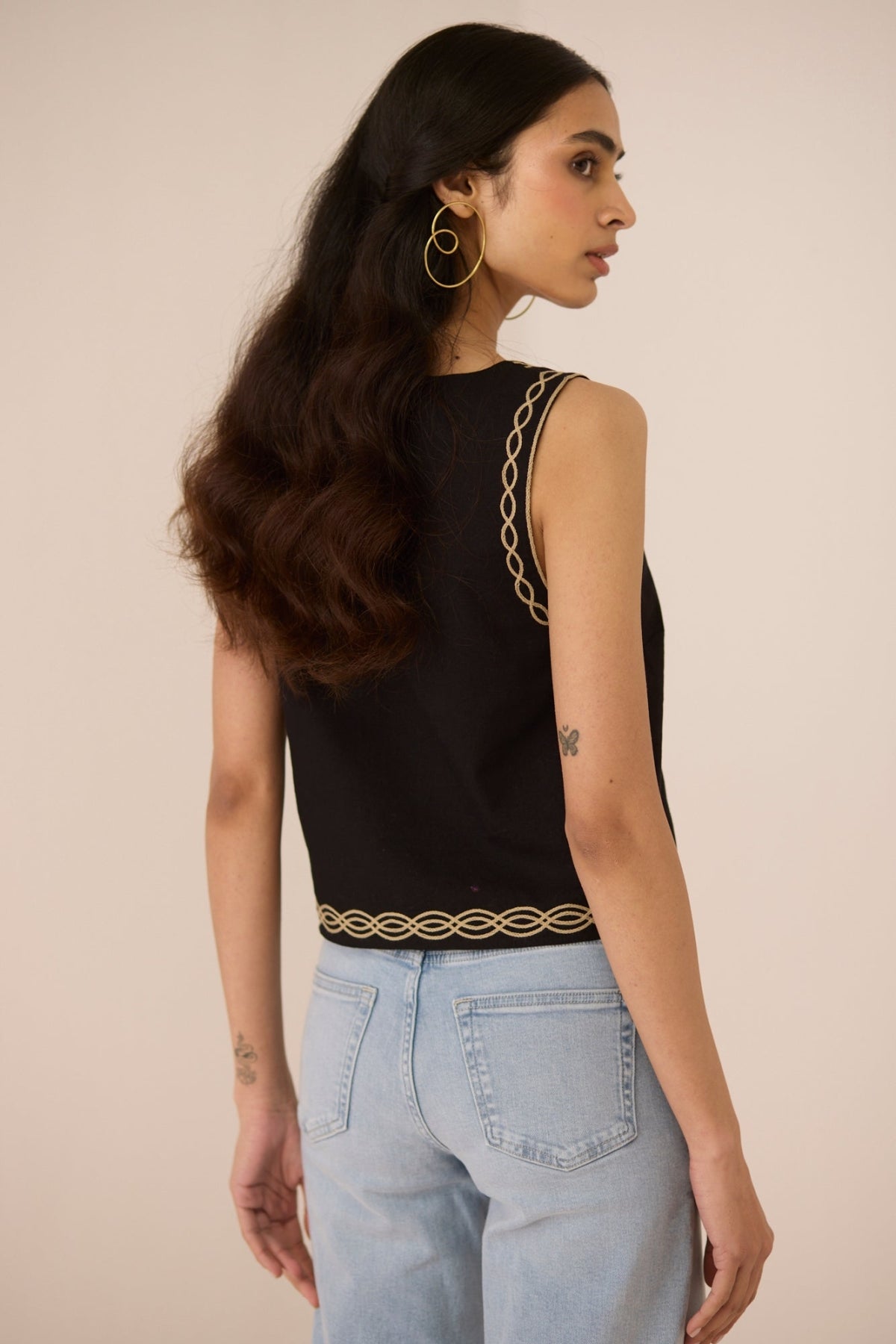 Woman wearing a black sleeveless top with gold detailing and light blue jeans on a beige background Calling June