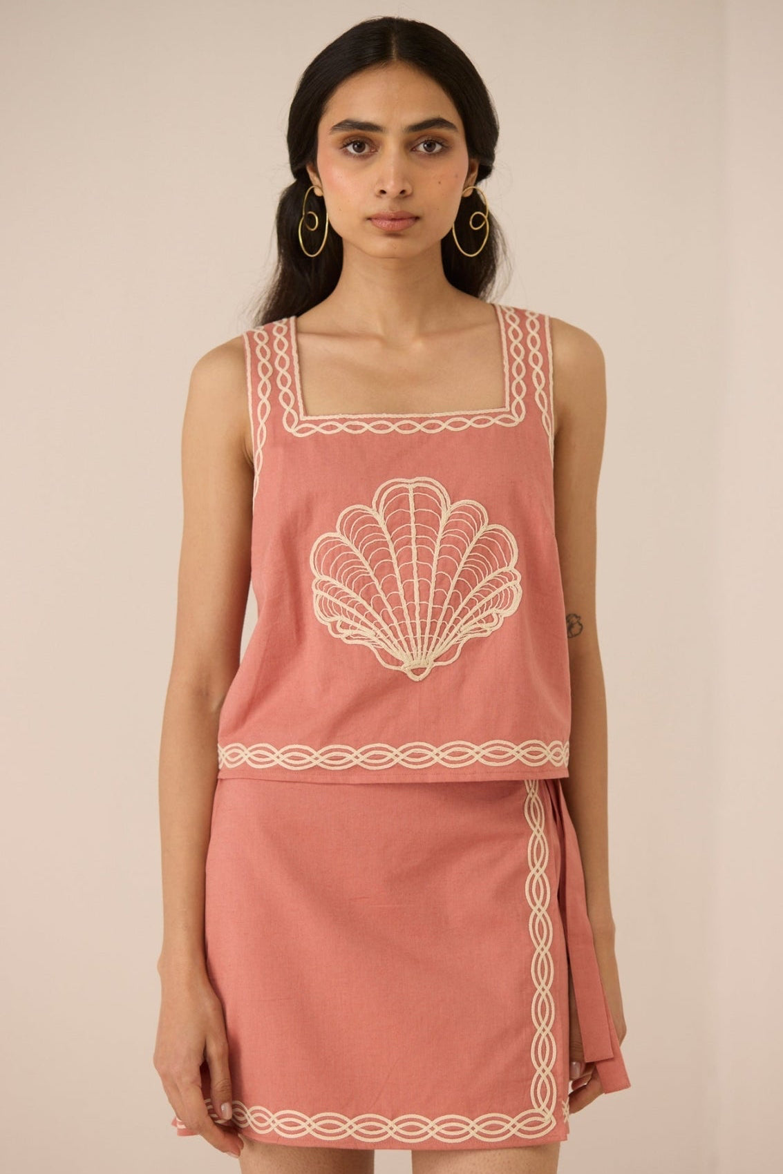 Woman wearing a pink sleeveless top and skirt with white embroidery on a beige background Calling June