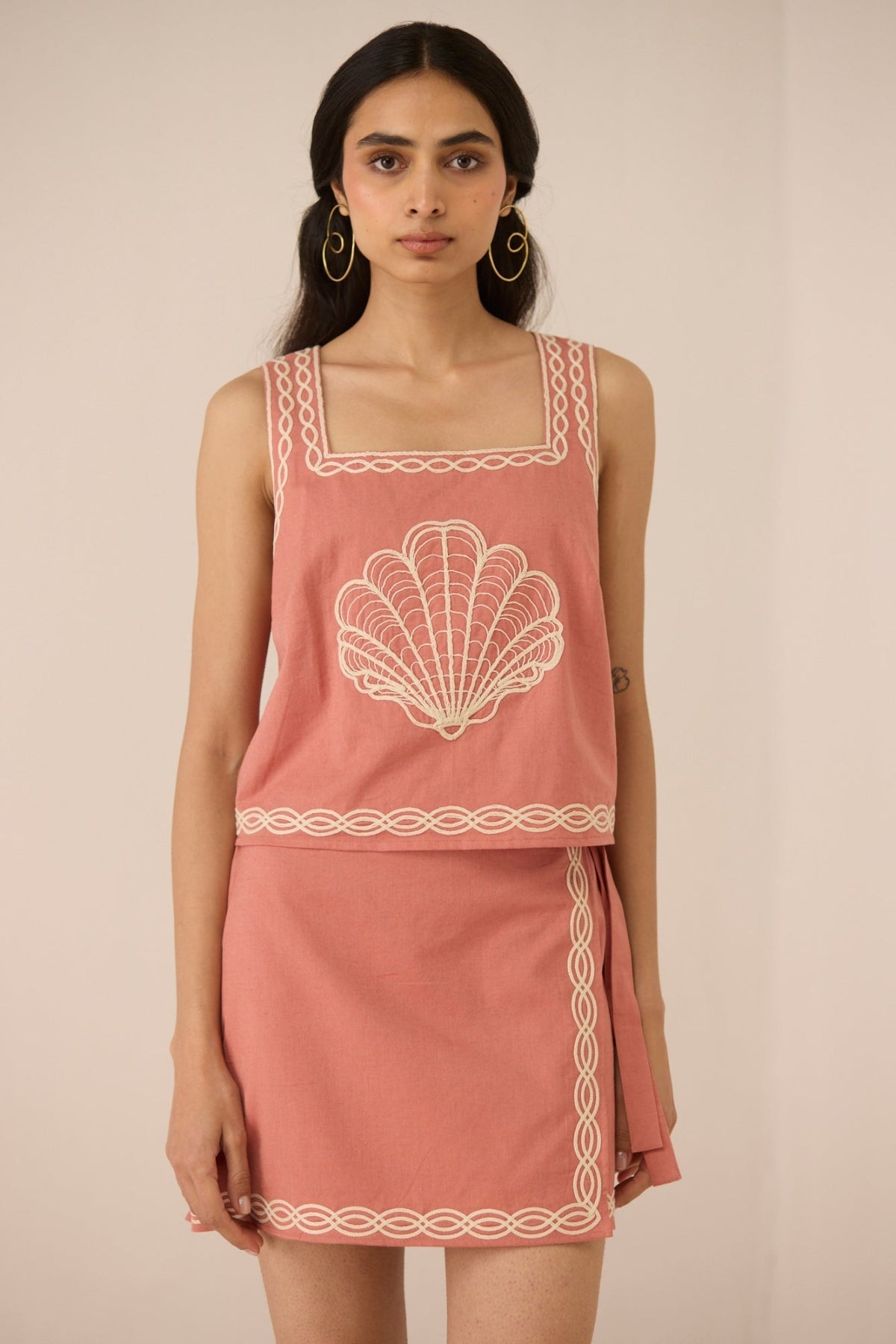 Woman wearing a pink sleeveless top and skirt with white embroidery on a beige background Calling June