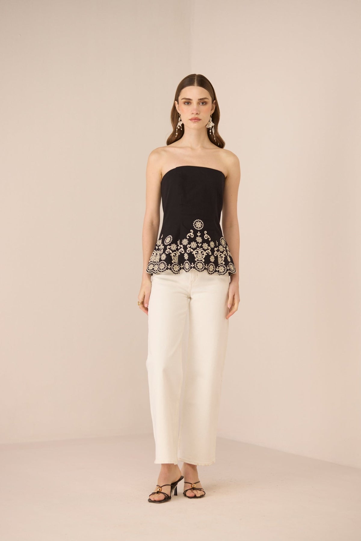 Woman wearing a black strapless top with white embroidery and white pants on a beige background Calling June