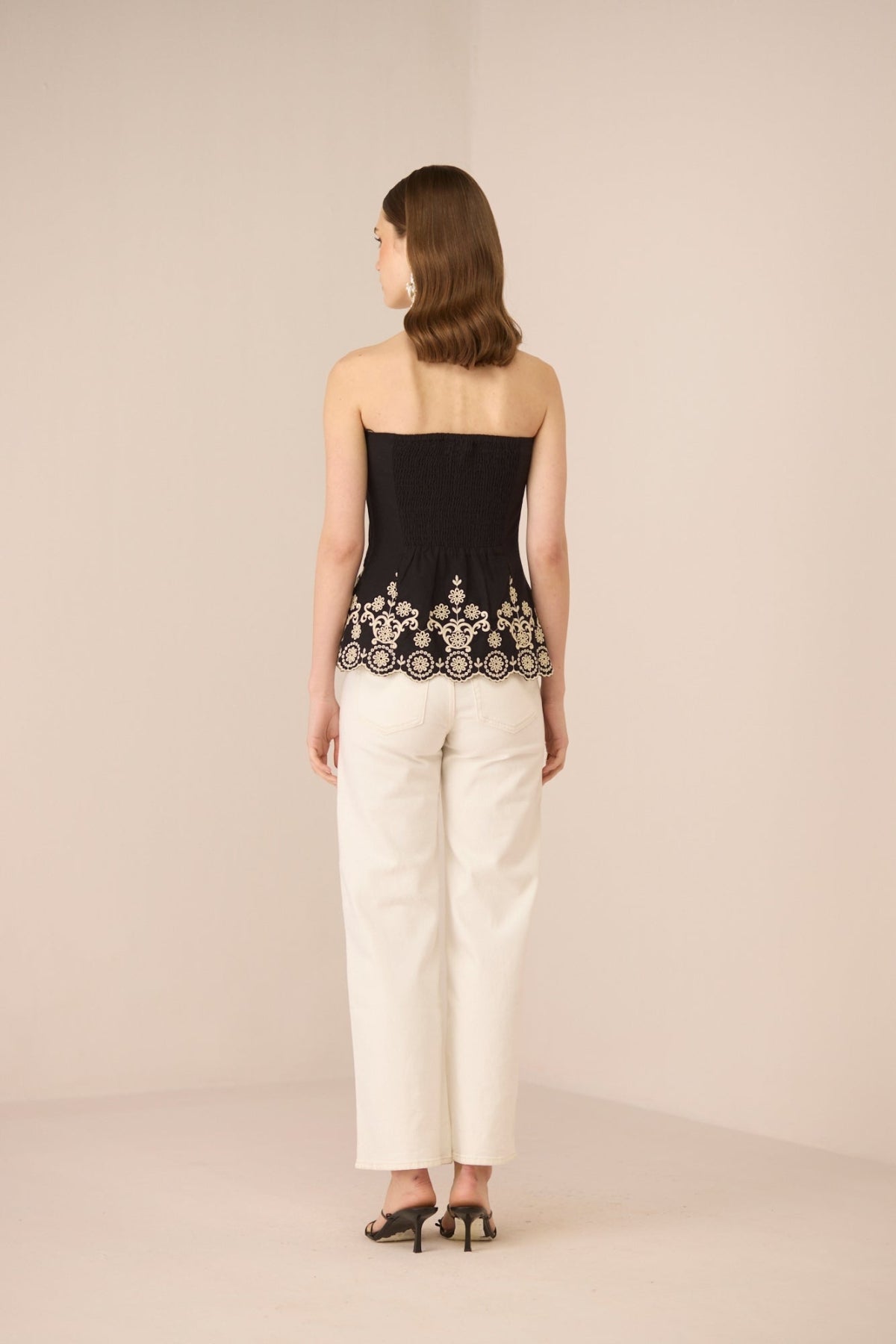 Woman wearing a black strapless top with lace detailing and white pants on a beige background Calling June