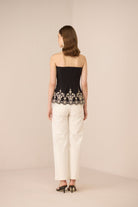 Woman wearing a black strapless top with lace detailing and white pants on a beige background Calling June