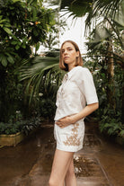 Woman in a white outfit with embroidery standing in a lush, green outdoor setting Calling June