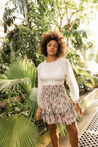 Woman in a white blouse and floral skirt standing in a tropical setting with greenery Calling June