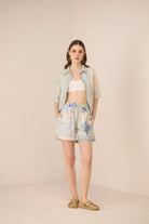 Woman wearing a light blue shirt, white top, and floral shorts on a beige background Calling June