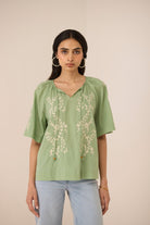 Woman wearing a green embroidered blouse with light blue jeans against a beige background Calling June