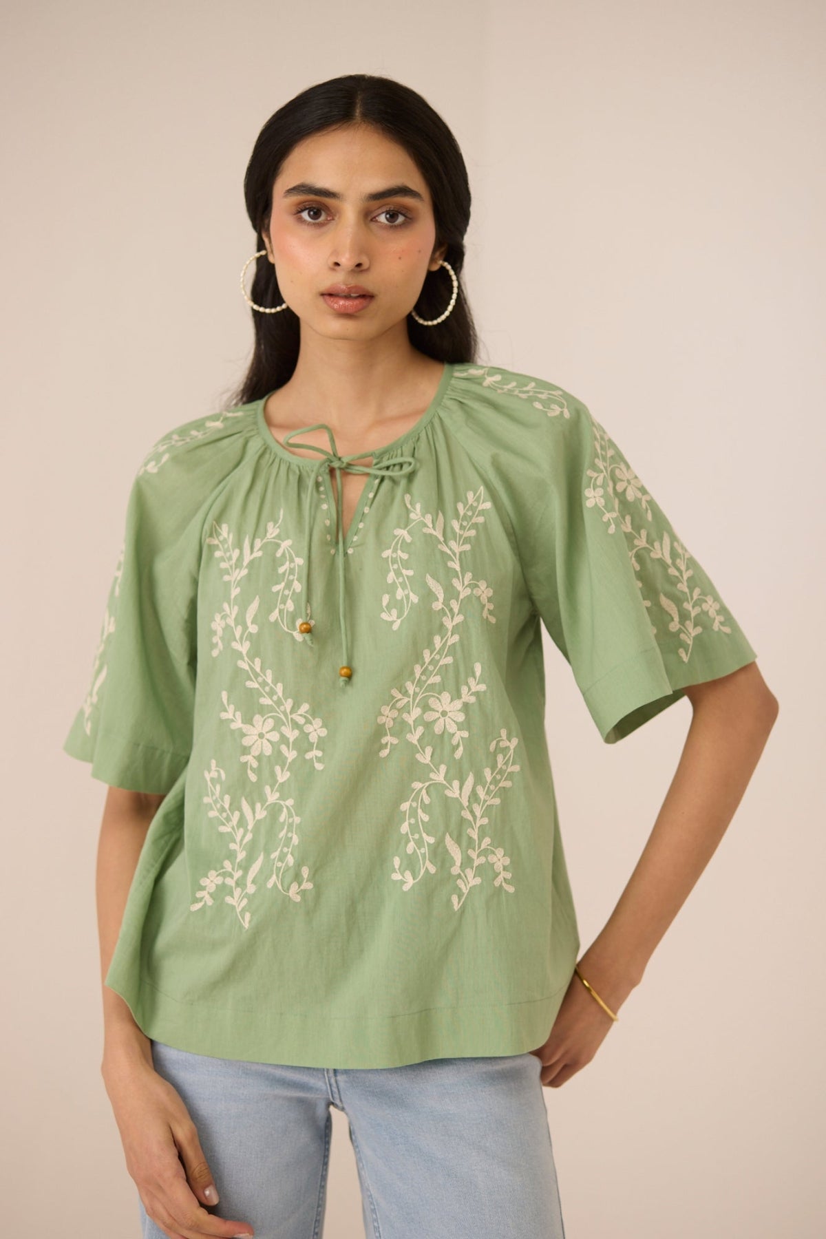 Woman wearing a green embroidered blouse with light blue jeans against a beige background Calling June