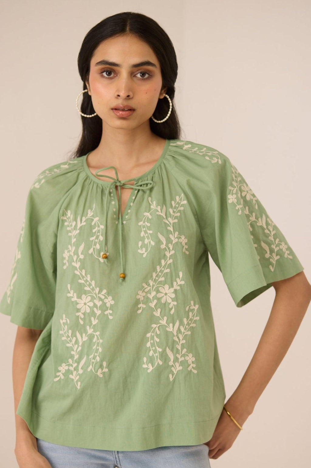 Woman wearing a green embroidered blouse with light blue jeans against a beige background Calling June