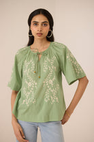 Woman wearing a green embroidered blouse with light blue jeans against a beige background Calling June