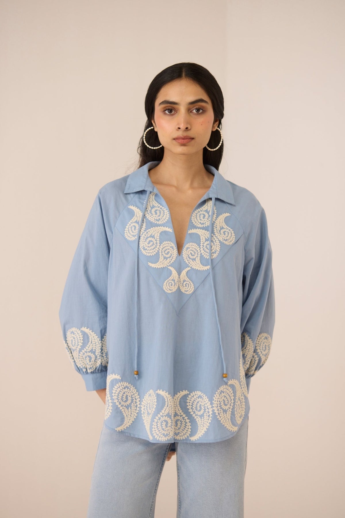 Woman wearing a light blue blouse with intricate white embroidery on a beige background Calling June