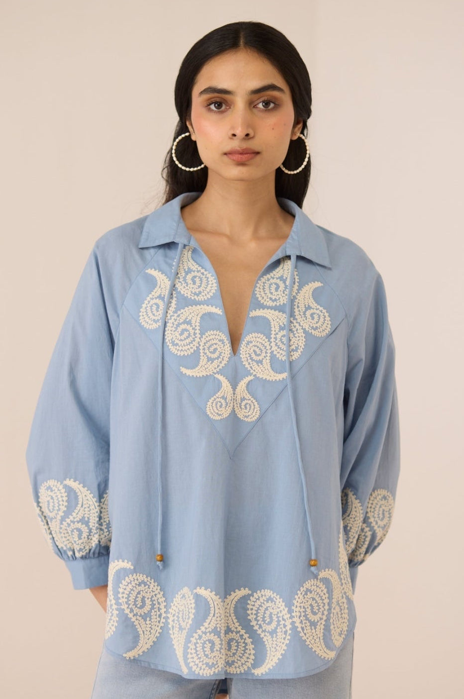 Woman wearing a light blue blouse with intricate white embroidery on a beige background Calling June