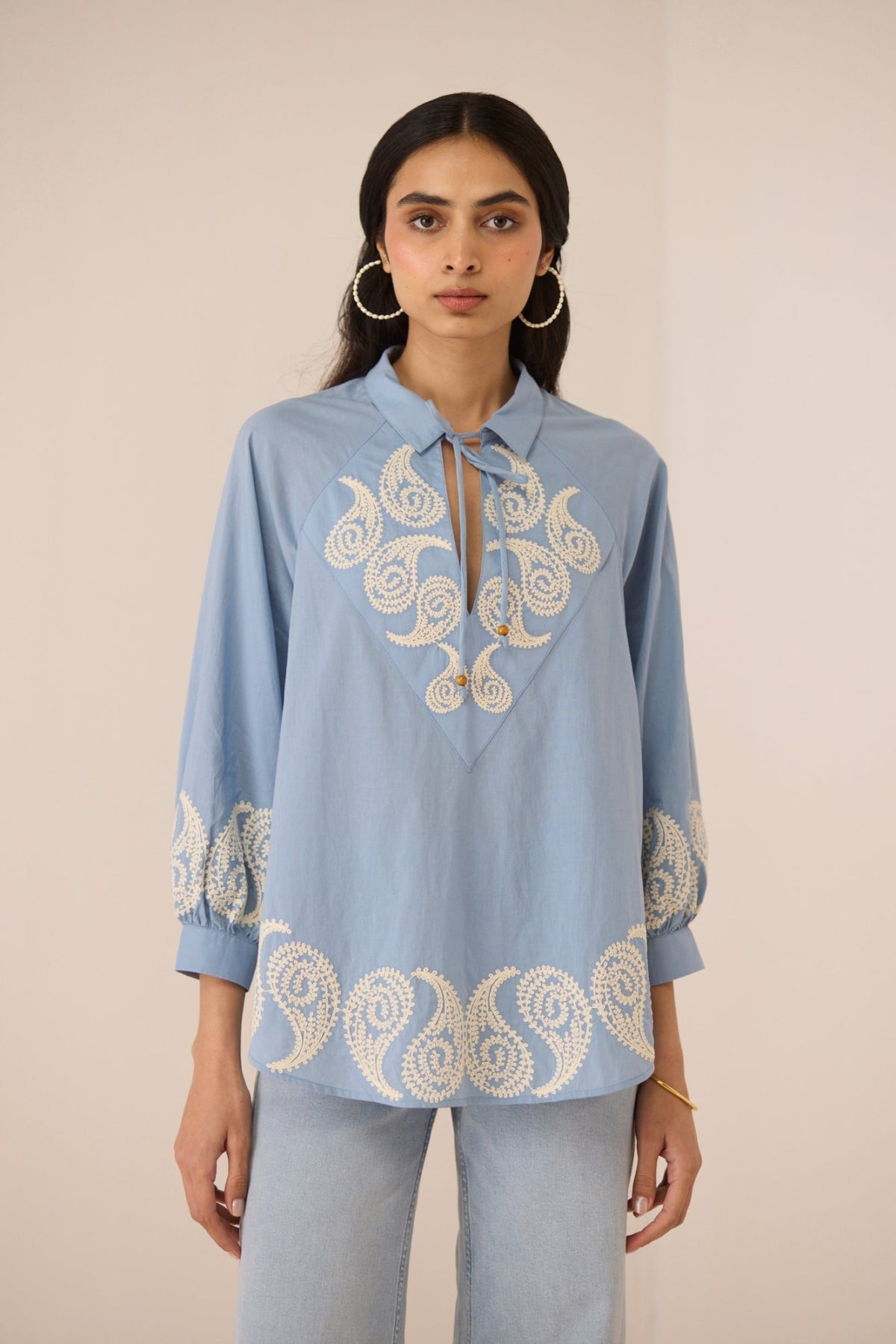 Woman wearing a light blue shirt with intricate white embroidery on a beige background Calling June