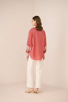 Woman wearing a pink blouse with floral embroidery and white pants on a beige background Calling June