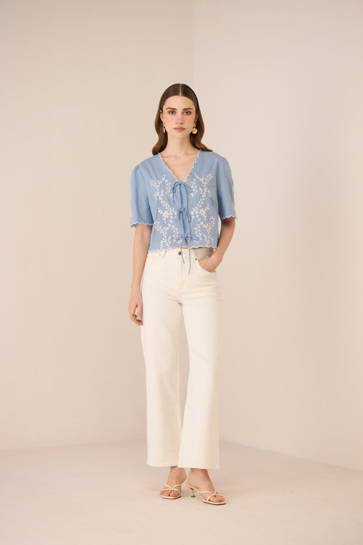 Woman wearing a blue embroidered blouse and white pants on a beige background Calling June
